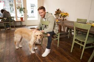 Lifestyle portrait of handsome smiling man sitting in pet friendly cafe with his beautiful dog, petting golden retriever, waiting for an order photo