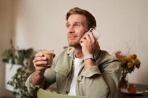imagen de sonriente, guapo joven rubio hombre con teléfono, Bebiendo café en cafetería, hablando a alguien terminado el teléfono, respondiendo un llamada foto
