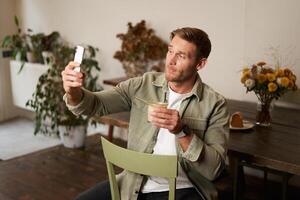 retrato de joven hermoso hombre toma slefie en su móvil teléfono aplicación, se sienta en café con vaso de café, destino en social medios de comunicación solicitud foto