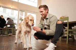Portrait of smiling handsome man with his dog, sitting on floor in cafe with golden retriever, giving a treat photo