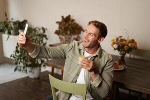 Portrait of handsome happy young man, taking selfie in cafe, posing with cup of coffee, chats using smartphone app, talks to a friend photo