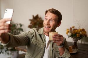 Portrait of smiling handsome young man, chats with friend from cafe, doing selfies with cup of coffee photo