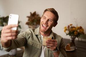 Close up portrait of happy, charismatic young blond man, taking selfie on smartphone, posing with cup of coffee, enjoys his drink in cafe, recommending a place to his followers online photo