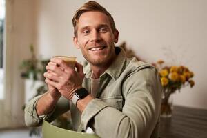 Portrait of good-looking young man with cup of coffee, sitting on chair in cafe, smiling and relaxing with his cappuccino drink photo