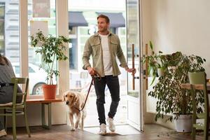 Portrait of handsome smiling young man, walking into the coffee shop with his cute dog, golden retriever on leash. Guy enters pet-friendly cafe photo