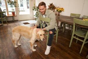 Close up portrait of handsome young man waiting for his order in coffee shop, petting a dog, resting in pet-friendly cafe photo