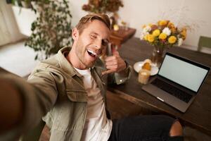 hermoso elegante joven hombre toma selfie en café mientras trabajando o estudiando remotamente con computadora portátil, muestra llamada teléfono mano firmar y sonriente a móvil teléfono cámara foto