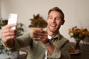 Cheerful handsome young man, taking selfie on mobile phone app, chats to someone and raising glass of coffee, cheers gesture, drinks cup of cappuccino and smiles at smartphone camera photo