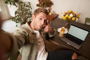 hermoso elegante joven hombre toma selfie en café mientras trabajando o estudiando remotamente con computadora portátil, muestra llamada teléfono mano firmar y sonriente a móvil teléfono cámara foto