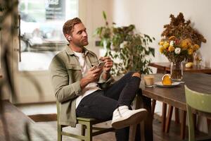 Portrait of young handsome man in casual clothes, sits in cafe and enjoys favourite song, listens to music in wireless headphones, vibing to the relaxing tunes in earphones, holding smartphone photo