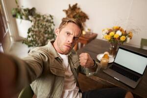 Portrait of serious man takes selfie with fist and threatening face, sits in cafe, poses in coffee shop in front of table with laptop and glass of coffee photo