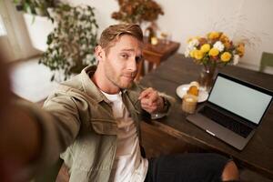 Handsome and confident young man taking selfie on mobile phone, pointing finger at camera, sitting in cafe with laptop, working from coffee shop, looking sassy photo