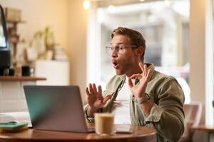 Portrait of man giving lessons online, shows okay, ok hand sign at laptop camera, talking to someone, having a meeting chat, sitting in cafe photo