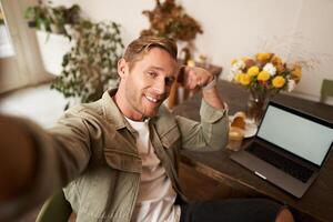 Portrait of handsome and confident man taking a selfie, flexing biceps, shows-off his muscles, sits in cafe in front of laptop, smiling at camera photo