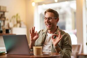 Portrait of young freelancer, digital nomad having an online meeting, chats, shows okay sign at laptop, confirm something, makes ok hand gesture, sits in cafe, works on remote photo