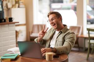 Happy smiling guy in glasses, sits in cafe, shows peace sign at laptop camera, chats, connects to online meeting photo