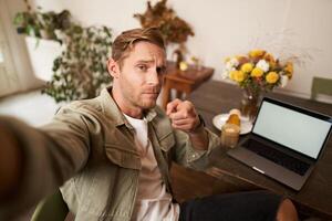 Confident, handsome man, pointing finger at his mobile phone, taking selfie, frowning sassy, sitting in cafe with laptop, drinking coffee photo