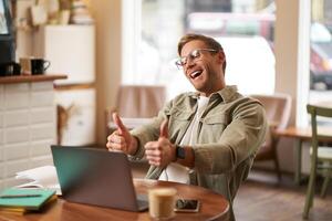 Portrait of satisfied, happy young man in glasses, working in cafe, sitting in co-working space with laptop, showing thumbs up, like and approve smth good, chatting, giving online lessons photo