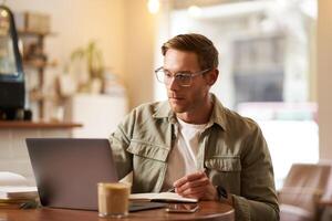 retrato de moderno hombre en anteojos, sentado en bastante vacío café con computadora portátil, escritura abajo notas en computadora portátil, estudiando en línea, remoto aprendizaje electrónico, paso web curso foto