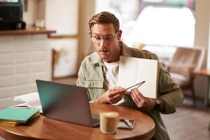 Portrait of young handsome man in glasses, private tutor teaching student online, pointing at his notebook, showing something, chats via laptop application, working remotely in cafe photo