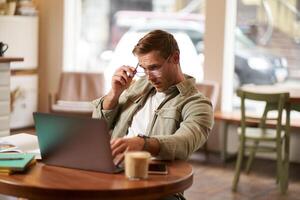 Portrait of a man working in cafe, looking surprised at camera, taking off glasses to stare puzzled at laptop screen, has a question about something on his computer. concept of remote jobs photo