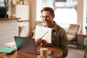 Portrait of young handsome man in glasses, private tutor teaching student online, pointing at his notebook, showing something, chats via laptop application, working remotely in cafe photo