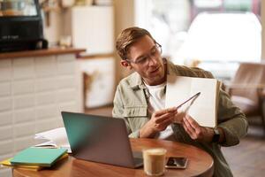Portrait of handsome young man giving lessons online, showing exercises in his notebook, looking at laptop, recording , sitting in cafe, quite co-working space photo