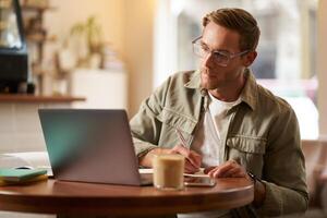 Portrait of handsome young guy in glasses, man studying, taking notes, looking at laptop screen during online lesson, attends a meeting and writing down information, sitting in cafe photo