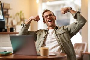 Portrait of handsome, pleased young man shows biceps, flexing muscles, sits in front of laptop in cafe, rejoicing, private coach having a chat with his client photo