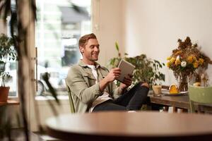 Portrait of happy, relaxed young man sitting in cafe alone, wearing wireless headphones, playing game on tablet, laughing and smiling, looking at his device. Concept of lifestyle and people photo