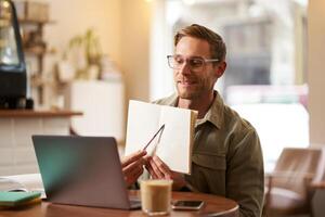 Portrait of young handsome man in glasses, private tutor teaching student online, pointing at his notebook, showing something, chats via laptop application, working remotely in cafe photo
