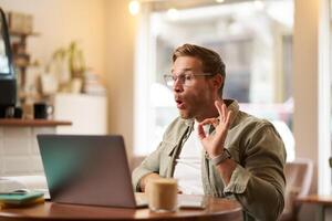 Portrait of young freelancer, digital nomad having an online meeting, chats, shows okay sign at laptop, confirm something, makes ok hand gesture, sits in cafe, works on remote photo