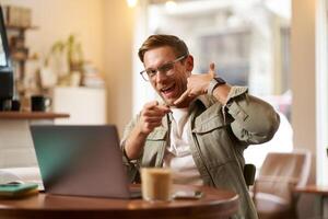 Portrait of cheerful, flirty guy in glasses, sitting with laptop computer in cafe, showing phone call hand gesture and winking at you, giving his number photo