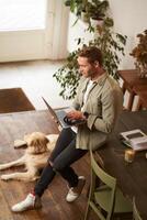 Vertical shot of handsome businessman, young man with laptop leans on table, sitting at co-working space with his dog. Young salesman checks his messages, smiling, working in an office with a pet photo