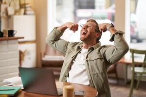 Portrait of pleased, smiling young man shouting with rejoice, sitting in cafe in front of laptop, flexing muscles, screaming from happiness, winning and celebrating photo