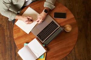 Top aerial view of male hands taking notes, writing in notebook, man sitting at round desk in cafe, working on laptop, studying photo