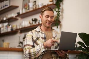 Image of young man in coffee shop, connects to call, talking online, chats with friends via laptop and smiles photo