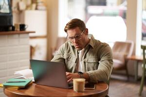 Portrait of angry man in glasses, looking frustrated and typing something on laptop, staring at screen with outraged face expression, sitting in cafe photo