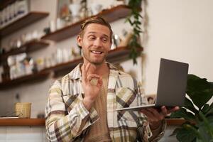 Portrait of satisfied smiling young man, holding laptop, showing okay, ok sign, approve smth good, satisfied by project result. Teacher giving online lesson in cafe, nod in approval photo