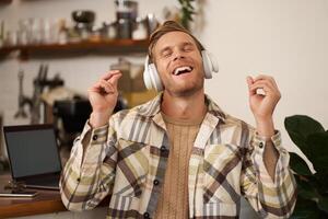 hermoso sonriente joven hombre, digital nómada, sentado en trabajo colaborativo abierto espacio, trabajando en café comercio, canto y escuchando a música en inalámbrico auriculares foto