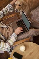 Vertical cropped photo of male hands and body, man petting the dog while working on laptop in cafe, drinking coffee