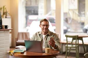 Portrait of handsome man in glasses, sits in cafe with laptop, smiles while reads a message on phone, checks his smartphone app, works from coffee shop photo