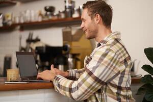 Portrait of handsome smiling man, sitting with laptop in a cafe, typing, working remotely from co-working space, drinking coffee, freelancing from public space photo