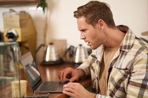 Portrait of man looks frustrated at laptop screen, staring at monitor with shocked face expression, reading message with frowned face, sitting in cafe, working remotely from coffee shop photo