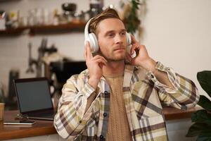 Portrait of handsome happy guy, freelancer working and listening to music in wireless headphones, dancing on his chair in cafe, enjoying favourite song in earphones photo