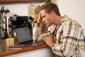 Image of adult man looking complicated at laptop screen, staring puzzled at his monitor, doing difficult task online, sitting in cafe, drinking coffee and working photo