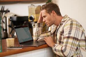 imagen de adulto hombre mirando Complicado a ordenador portátil pantalla, curioso perplejo a su monitor, haciendo difícil tarea en línea, sentado en cafetería, Bebiendo café y trabajando foto