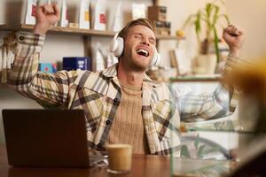 retrato de entusiasmado, contento joven hombre sentado detrás el mostrador en cafetería, trabajando en computadora portátil, Bebiendo café, escuchando música en inalámbrico auriculares, canto alto Nota foto