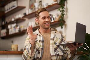 Image of young man in coffee shop, connects to call, talking online, chats with friends via laptop and waves hand at camera to say hi photo