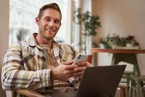 Portrait of young good-looking man, cafe visitor, sitting with laptop and smartphone, smiling at camera, freelancer working remotely from co-working space photo